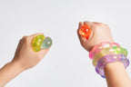Two hands holding and wearing translucent, glittery sensory toys against a plain light gray background. One hand holds a yellow-green and blue glitter toy, and the other wrist is stacked with pink, green, purple, and orange glitter bracelets.