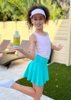 Young girl in a white visor and turquoise skirt happily holding a bottle of California Baby - Calendula™ Shampoo + Conditioner Bundle outdoors, showcasing cozy baby essentials.