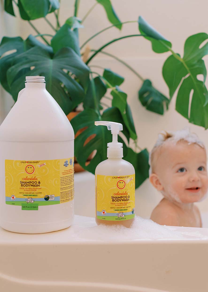 California Baby - Calendula™ Shampoo & Bodywash bottles next to a bubbly baby in a bathtub with leafy green plants in the background, showcasing a cozy and natural bath time setting.
