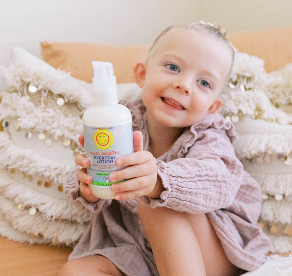 Young child sitting on a cozy couch holding a bottle of California Baby - (No Fragrance) Super Sensitive™ Everyday Lotion, surrounded by soft neutral pillows in a stylish children’s boutique setting.