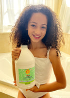 A smiling young girl with curly hair holding a large bottle of California Baby - Calming™ Hair Conditioner in a bright bathroom, showcasing gentle and soothing hair care.
