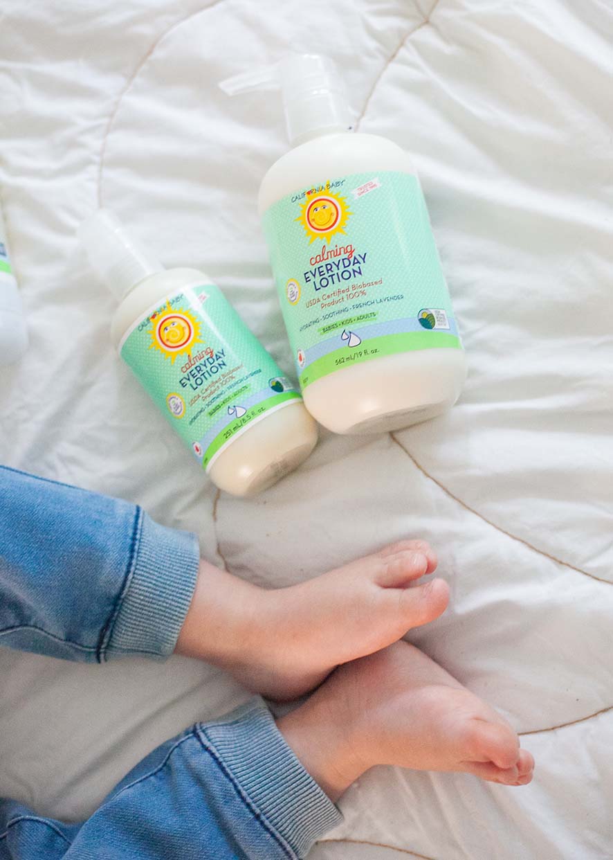 Two bottles of California Baby - Calming™ Everyday Lotion on a soft white blanket near a child’s bare feet wearing blue pants, highlighting cozy baby essentials and mom life essentials.