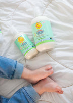 Two bottles of California Baby - Calming™ Everyday Lotion on a soft white blanket near a child’s bare feet wearing blue pants, highlighting cozy baby essentials and mom life essentials.