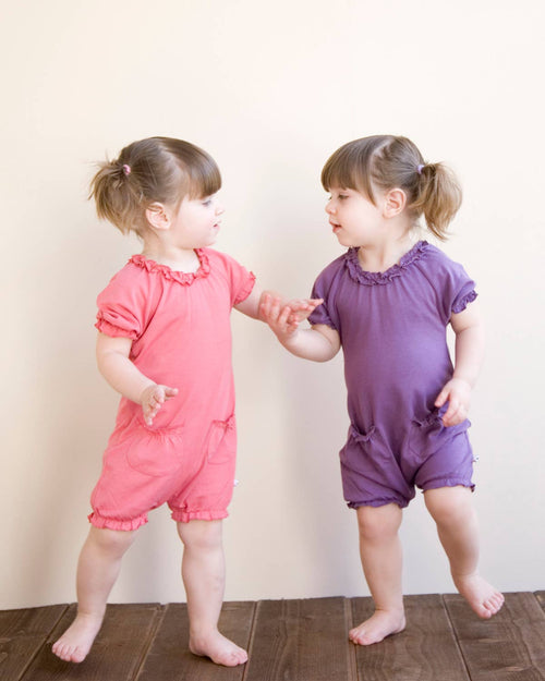 Two toddlers wearing colorful rompers standing on a wooden floor against a light wall, featuring babysoy Inc - Basic Bubble Romper: Pink Lemon in a soft, breathable fabric ideal for cozy baby essentials and stylish kids clothing.
