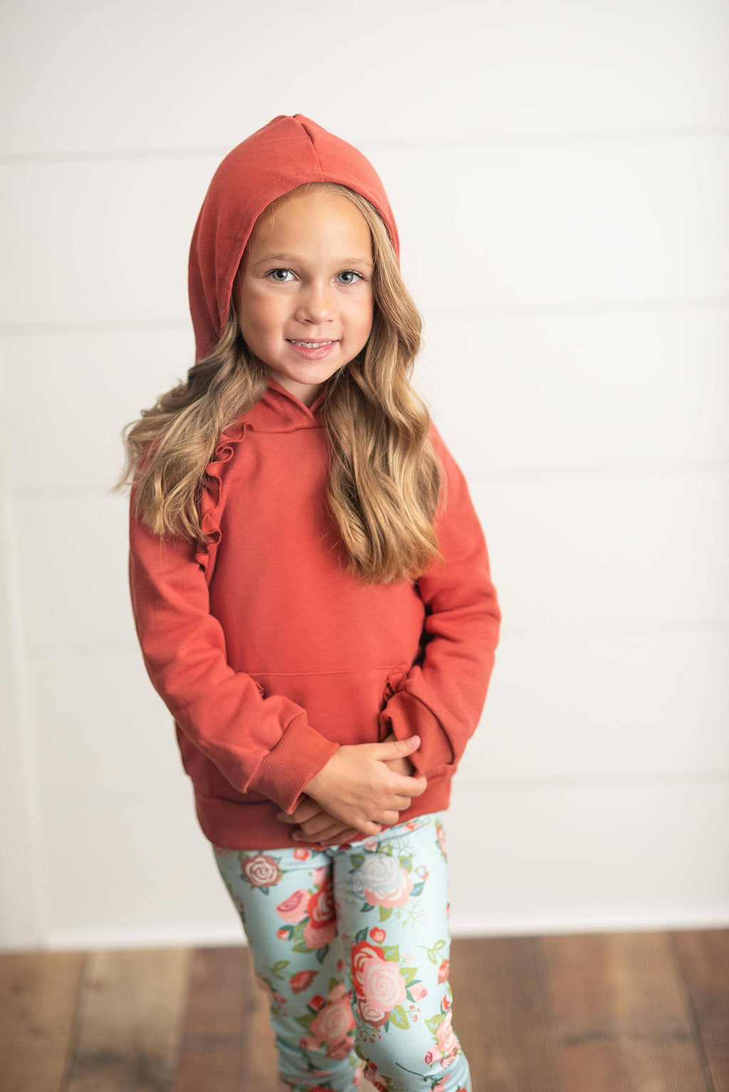 A young girl with blonde hair wearing a rust-orange hooded sweatshirt with ruffle details on the sleeves and light blue floral leggings, standing against a white paneled wall.