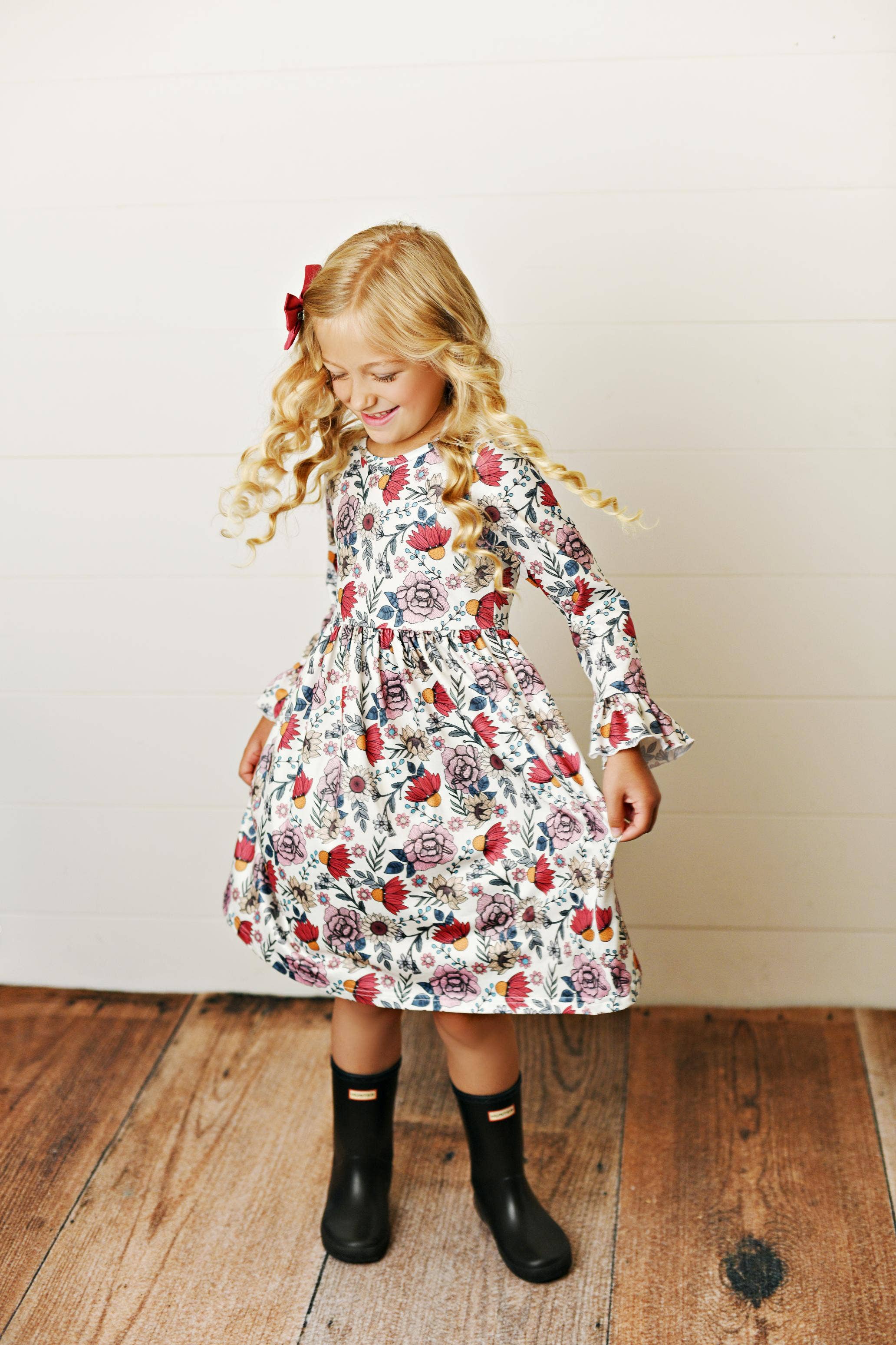 Young girl wearing a floral dress standing on a wooden floor against a white wall.