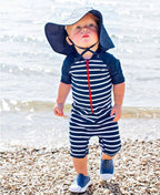 Toddler wearing a navy striped swim romper and a RuffleButts + RuggedButts - Kids Sun Protective Hat while standing on a rocky beach, showcasing stylish kids clothing and sun safety.