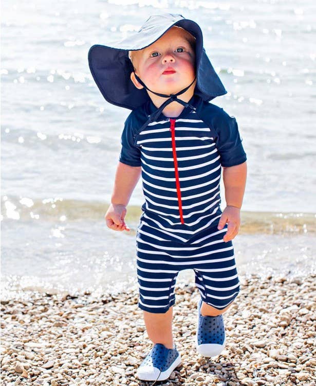 Toddler wearing a navy striped swim romper and a RuffleButts + RuggedButts - Kids Sun Protective Hat while standing on a rocky beach, showcasing stylish kids clothing and sun safety.
