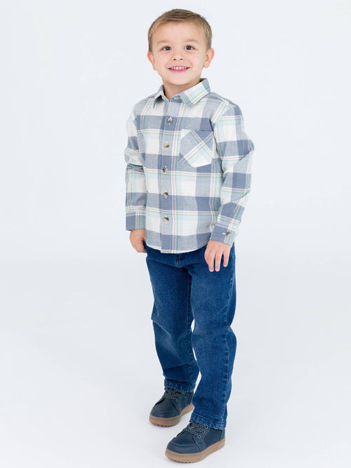 Young boy smiling and standing wearing the RuffleButts + RuggedButts - Boys Alpine Plaid Flannel Long Sleeve Button Down Shirt: Blue paired with dark jeans and navy shoes in a bright studio setting from SophistiKids Boutique.