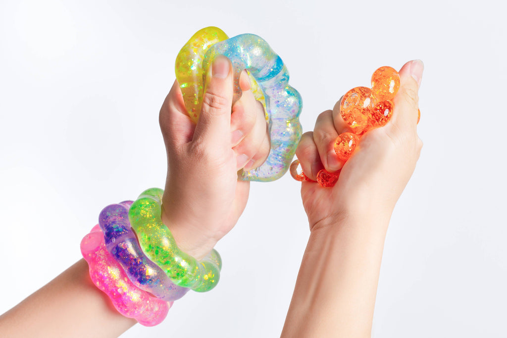 Two hands squeezing translucent glittery stress balls in yellow, blue, orange, green, and pink, with matching wristbands on a plain white background.