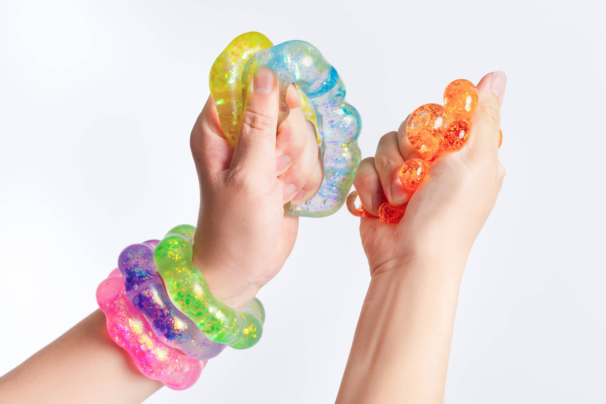 Two hands squeezing translucent glittery stress balls in yellow, blue, orange, green, and pink, with matching wristbands on a plain white background.