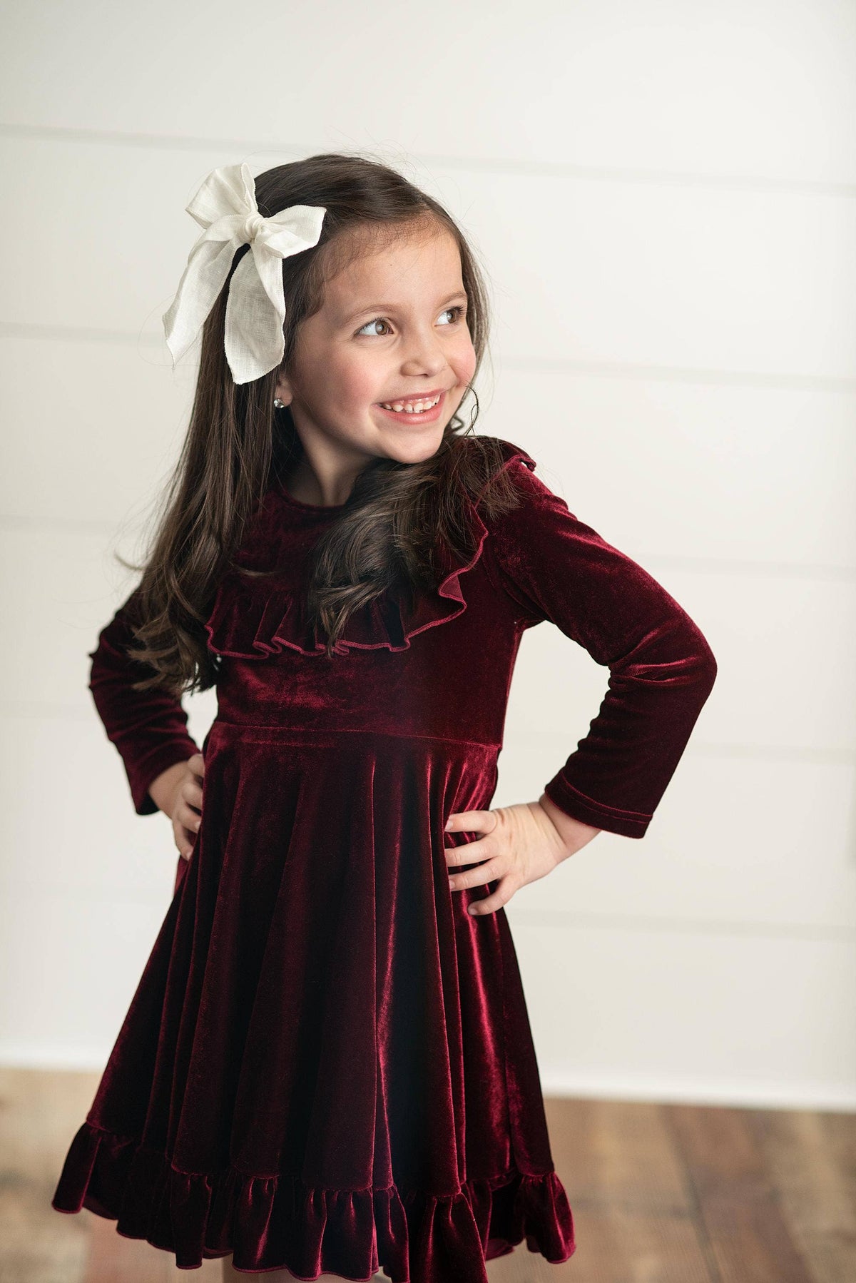 Young girl wearing a burgundy velvet dress with a white bow in her hair against a white background.