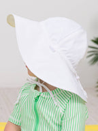 A toddler wearing a white RuffleButts + RuggedButts - Kids Sun Protective Hat with a wide brim, paired with a green striped outfit, in a bright and cozy setting.