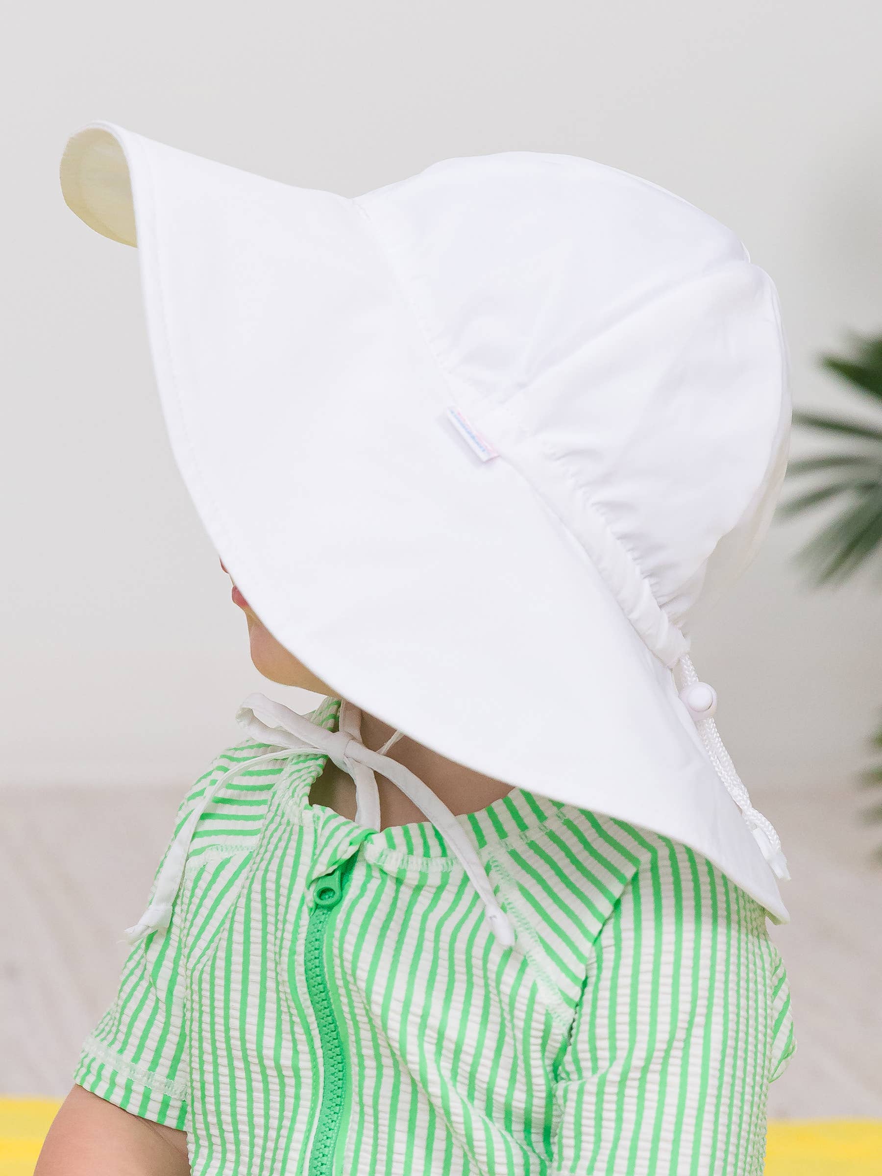 A toddler wearing a white RuffleButts + RuggedButts - Kids Sun Protective Hat with a wide brim, paired with a green striped outfit, in a bright and cozy setting.