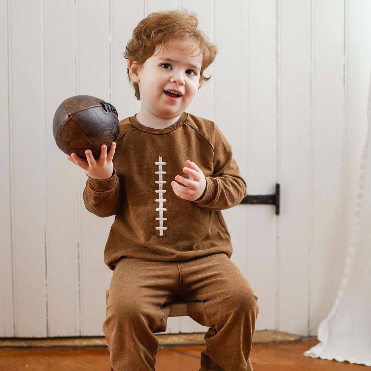 Child wearing a brown outfit with a football design, holding a vintage-style football.