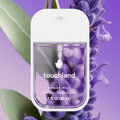 Close-up of Touchland - Pure Lavender Power Mist Hydrating Hand Sanitizer bottle with clear liquid, set against lavender flowers and soft green leaves for a calming, natural effect.