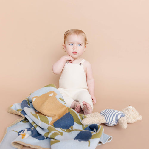 Baby seated on a soft beige surface surrounded by The Blueberry Hill - Blanket Blueberry Bear Bee Organic Cotton Spring Baby Decor featuring nature-inspired colors and playful bear and bee designs.