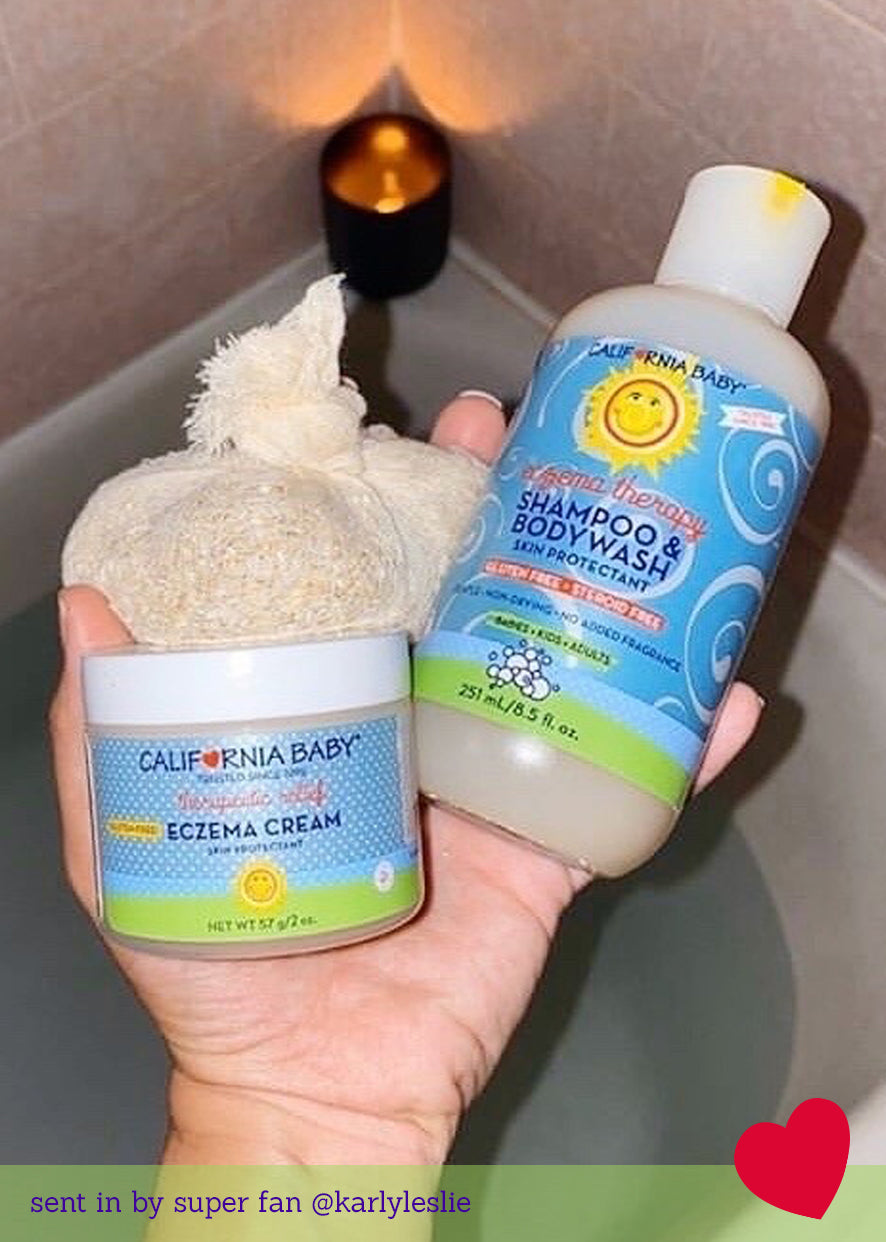 Hand holding California Baby - Therapeutic Relief™ Eczema Shampoo & Bodywash and eczema cream next to a bath sponge, with a lit candle in the background, showcasing soothing wellness products for babies.