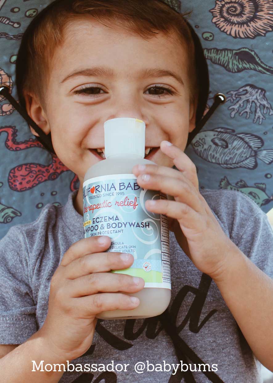 Smiling toddler holding a bottle of California Baby - Therapeutic Relief™ Eczema Shampoo & Bodywash, featured by a curated children’s boutique specializing in stylish kids clothing and mom life essentials.