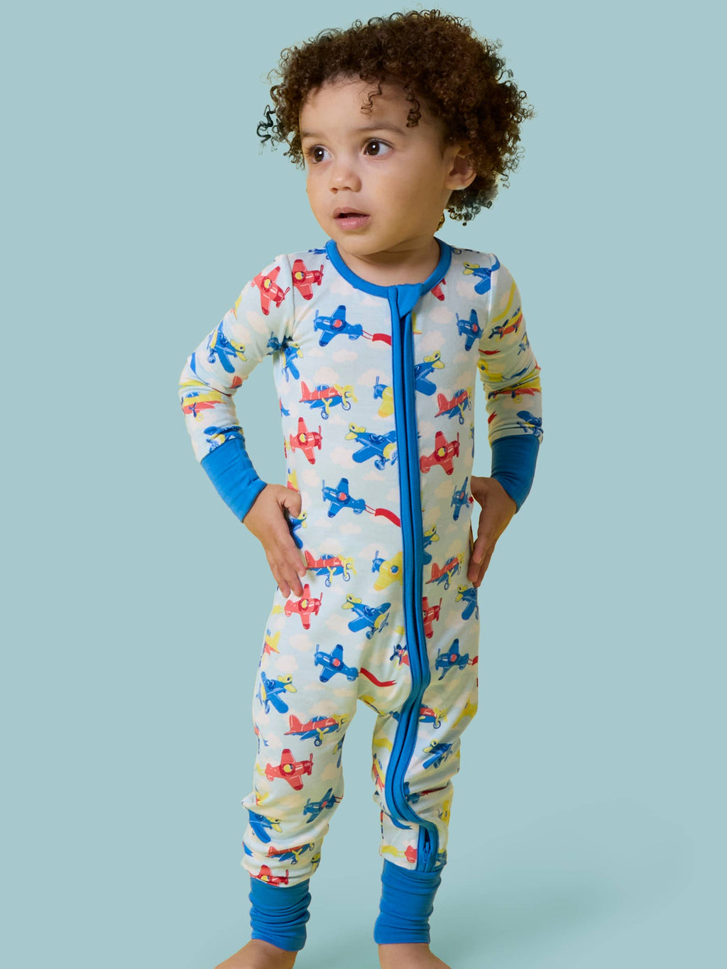 Toddler wearing RuffleButts + RuggedButts - SoftSnooze™ Baby Bamboo Viscose Plane Parade Convertible One Piece Footie Pajama with colorful airplane print and blue trim, standing against a light blue background in a children’s boutique style.