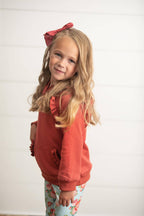 A young girl with blonde hair wearing a rust-orange ruffle-trimmed hoodie and light blue floral leggings, standing against a white paneled wall.