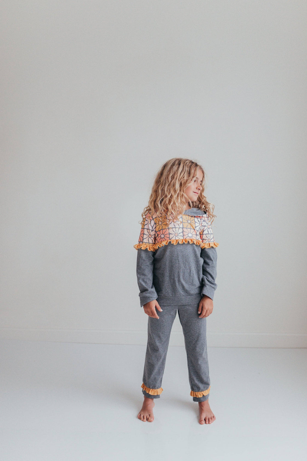 A young girl with long curly blonde hair stands barefoot in a studio setting, wearing a heather gray two-piece pajama set with a colorful patterned yoke and orange pom-pom trim.