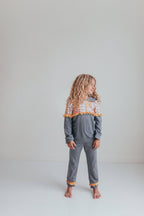A young girl with long curly blonde hair stands barefoot in a studio setting, wearing a heather gray two-piece pajama set with a colorful patterned yoke and orange pom-pom trim.