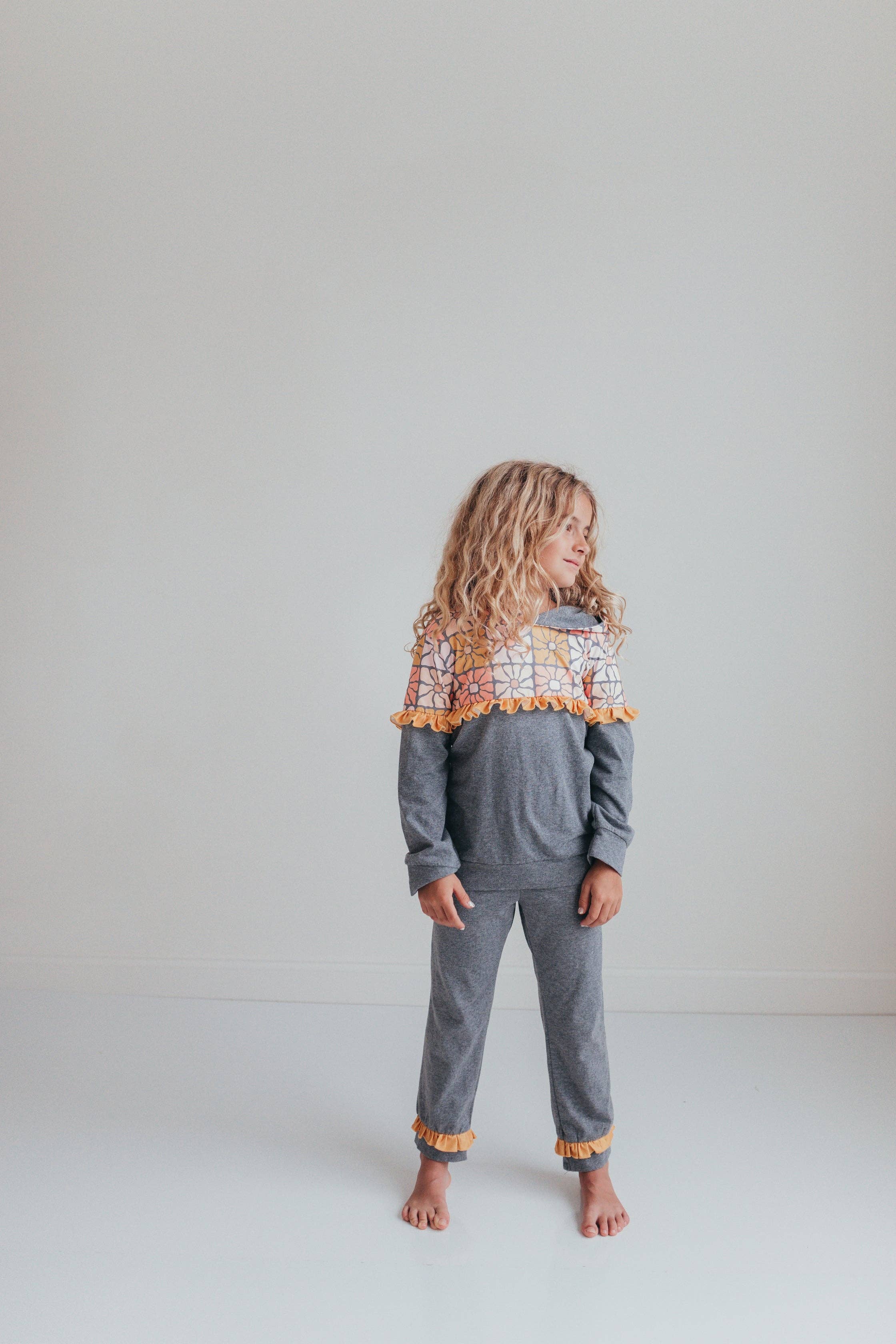 A young girl with long curly blonde hair stands barefoot in a studio setting, wearing a heather gray two-piece pajama set with a colorful patterned yoke and orange pom-pom trim.