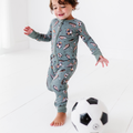 Toddler in playful green footie pajamas with soccer ball and sneaker prints, smiling while kicking a black and white soccer ball, showcasing Kiki + Lulu - Goal Hard or Go Home Convertible Footies.
