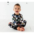 Toddler smiling and sitting cross-legged wearing Kiki + Lulu - Racecar Convertible Footies with colorful racecar patterns on a black and white checkered background, perfect for stylish kids clothing and cozy baby essentials.