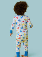 Toddler wearing RuffleButts + RuggedButts - SoftSnooze™ Baby Bamboo Viscose Plane Parade Convertible One Piece Footie Pajama with colorful airplane prints, standing against a light blue background.
