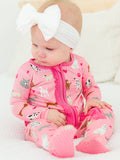 Baby wearing RuffleButts + RuggedButts - SoftSnooze™ Baby Girls Bamboo Viscose Pink Puppy Playtime Footed Ruffle One Piece Pajama: Pink, sitting on a white surface with a large white bow headband.