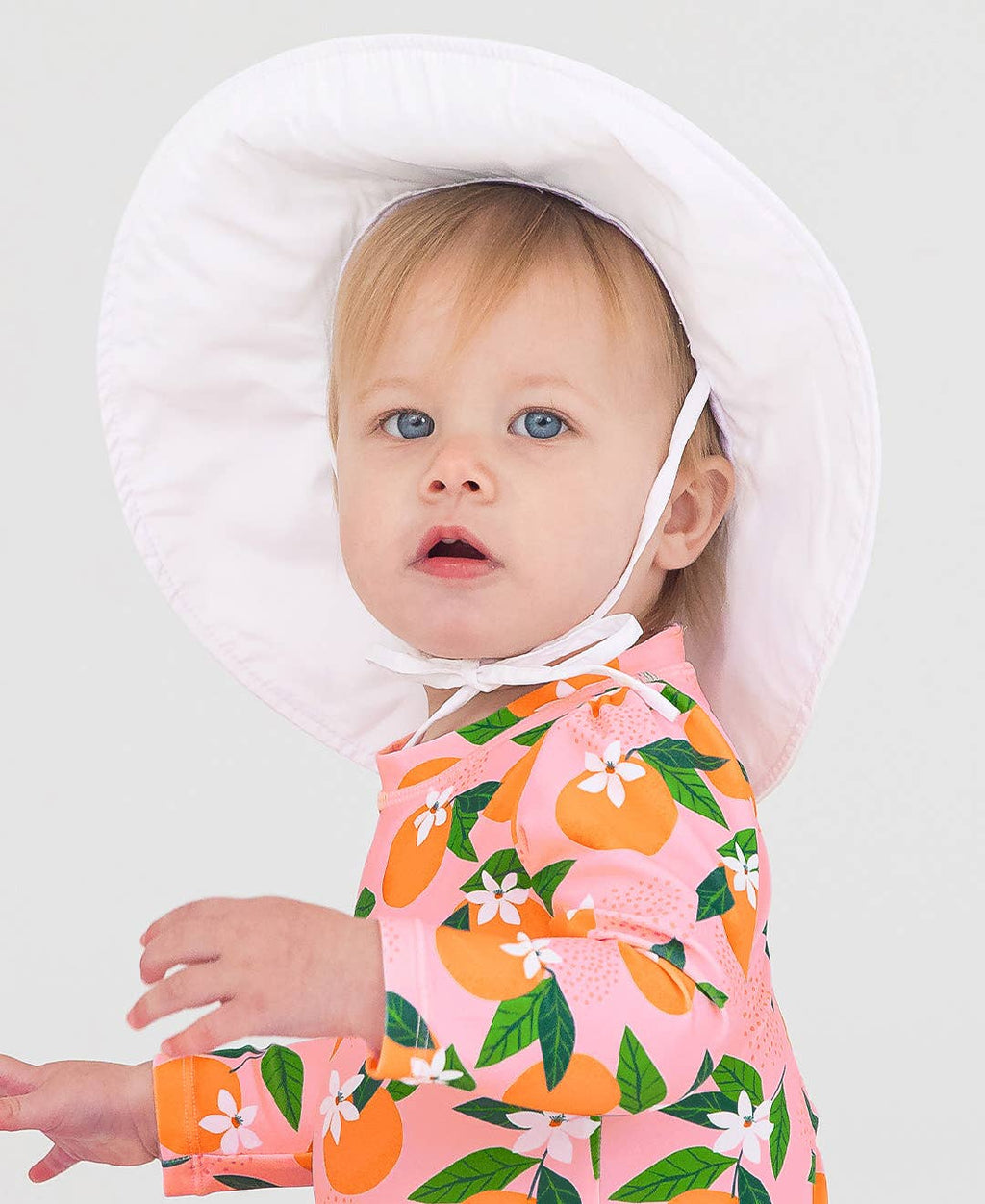 Toddler wearing a RuffleButts + RuggedButts - Kids Sun Protective Hat in white, paired with a pink long-sleeve outfit with orange and green fruit patterns, showcasing stylish kids clothing.