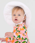 Toddler wearing a RuffleButts + RuggedButts - Kids Sun Protective Hat in white, paired with a pink long-sleeve outfit with orange and green fruit patterns, showcasing stylish kids clothing.