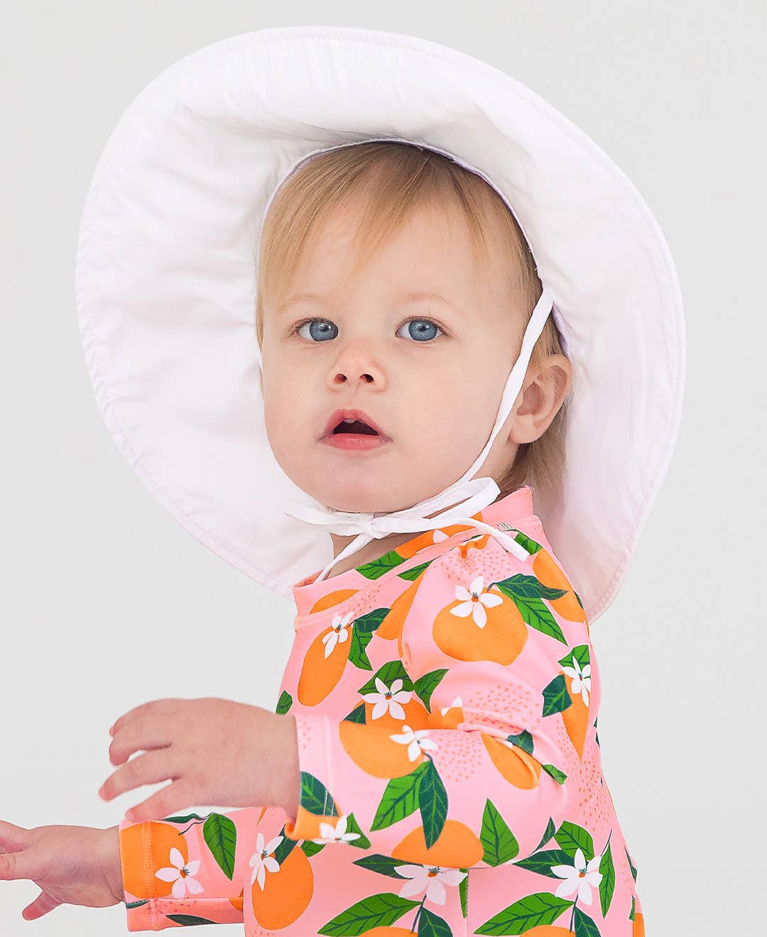 Toddler wearing a RuffleButts + RuggedButts - Kids Sun Protective Hat in white, paired with a pink long-sleeve outfit with orange and green fruit patterns, showcasing stylish kids clothing.