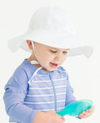 Toddler wearing a white RuffleButts + RuggedButts - Kids Sun Protective Hat and a blue striped rash guard while playing with a green shovel, showcasing stylish kids clothing from a curated children’s boutique.