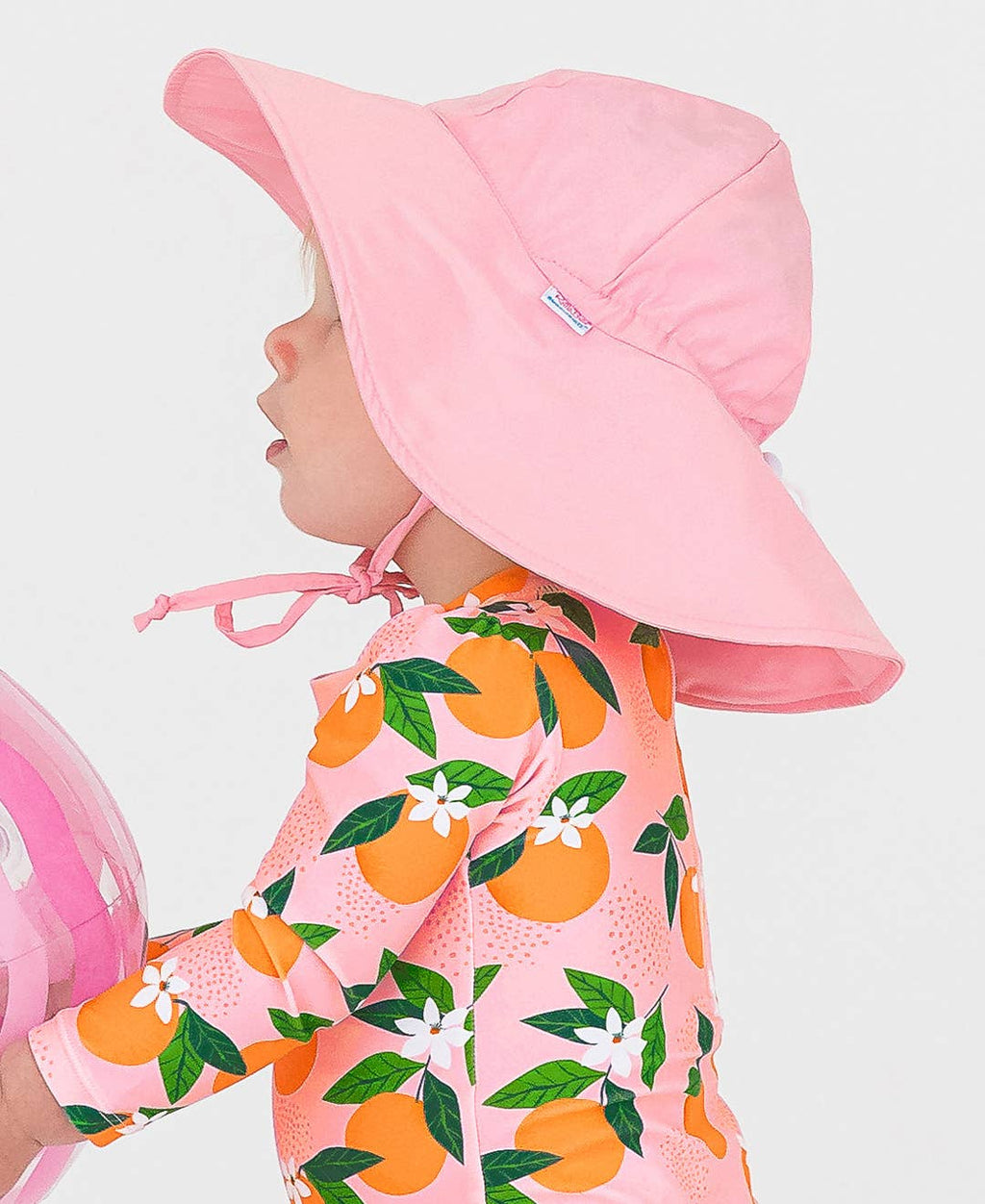 Toddler wearing a pink RuffleButts + RuggedButts - Kids Sun Protective Hat paired with a vibrant orange and green patterned outfit, showcasing stylish kids clothing from a curated children’s boutique.