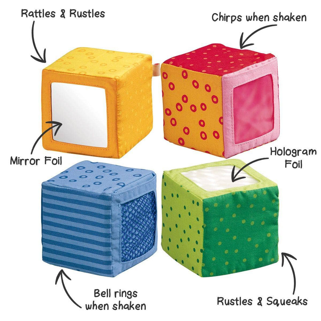 Four colorful soft fabric blocks with interactive features including mirror foil, chirping sound, bell rings, rustles, squeaks, and hologram foil for sensory play in the Happy Quartett Baby's 4 Piece Soft Block Set.