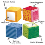 Four colorful soft fabric blocks with interactive features including mirror foil, chirping sound, bell rings, rustles, squeaks, and hologram foil for sensory play in the Happy Quartett Baby's 4 Piece Soft Block Set.