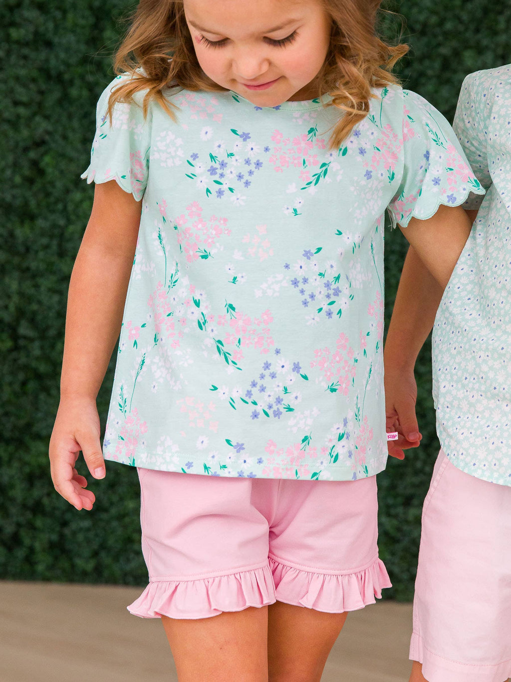 Young girl wearing RuffleButts + RuggedButts - Girls Pink Knit Ruffle Trim Shorts: Pink paired with a pastel floral top, standing against a dark green leafy background in a children's boutique setting.