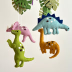 Colorful felt dinosaurs and green leaves suspended in a whimsical nursery decor piece, featuring the Mobile - Felt Baby - Jurassic Dinosaur, perfect for a curated children’s boutique.