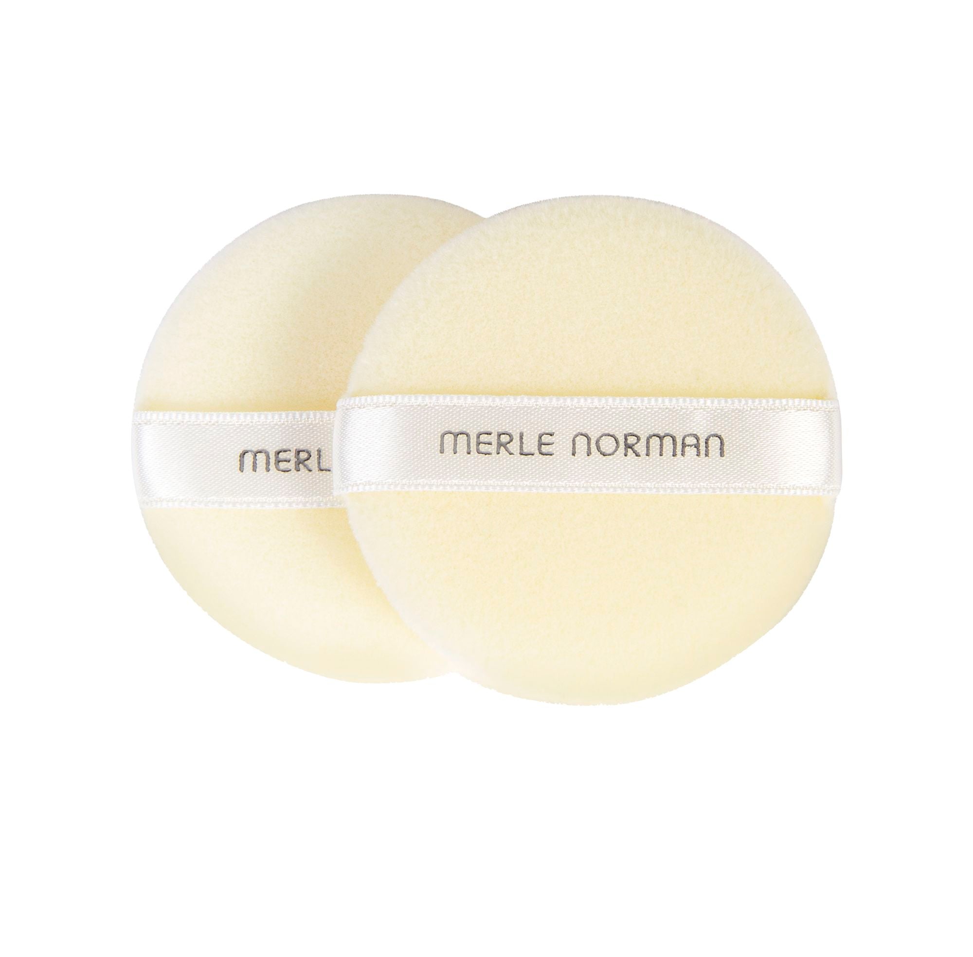 Two soft, round, beige powder puffs with satin bands labeled Merle Norman, designed for smooth makeup application. Merle Norman Cosmetics - Loose Powder Puffs.