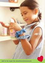 Young child wearing blue scrunchies holding and applying California Baby - Overtired & Cranky™ Everyday Lotion with a small amount on her arm, showcasing cozy baby essentials in a warm, soft-lit setting.
