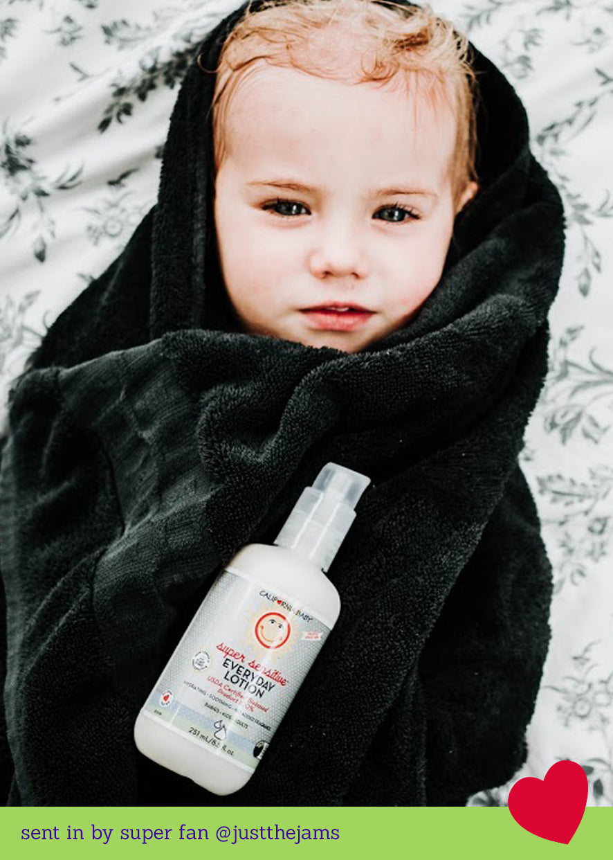 Child wrapped in a soft black towel holding a bottle of California Baby - (No Fragrance) Super Sensitive™ Everyday Lotion, highlighting cozy baby essentials for sensitive skin.