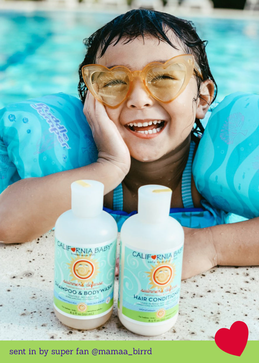 Smiling child wearing heart-shaped glasses and blue floaties in a pool with California Baby - Swimmer's Defense™ Shampoo & Bodywash bottles at the edge, showcasing mom life essentials.