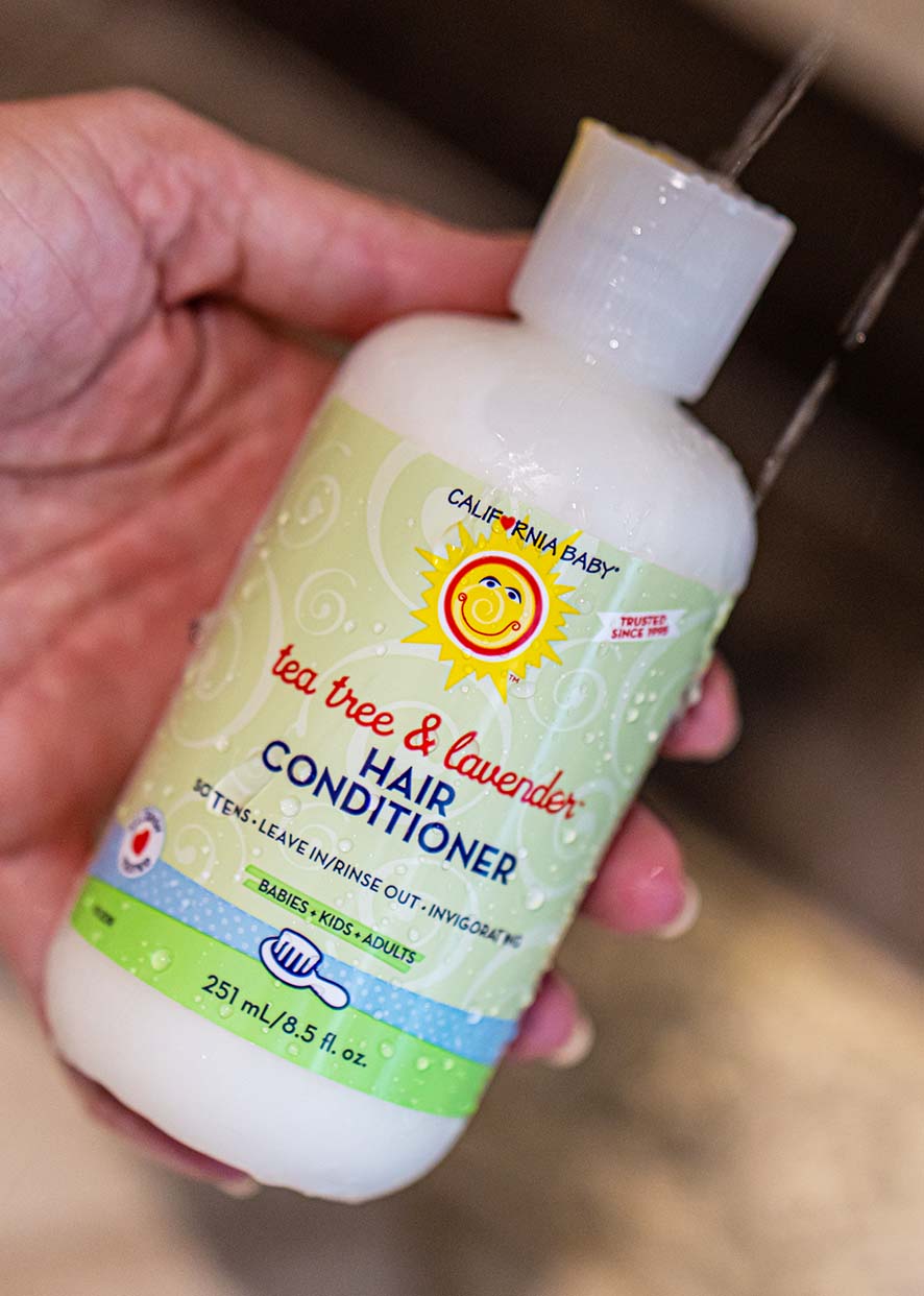 Hand holding a bottle of California Baby - Tea Tree & Lavender Hair Conditioner with water pouring over it, showcasing a soothing conditioner suitable for babies, kids, and adults.
