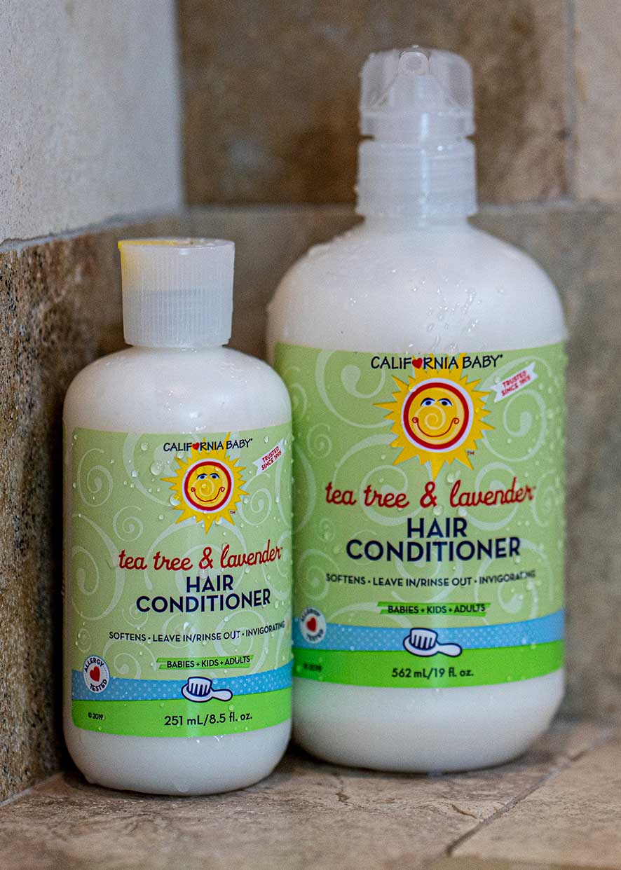 Two bottles of California Baby - Tea Tree & Lavender Hair Conditioner with green labels, designed for babies, kids, and adults, displayed in a bathroom setting.