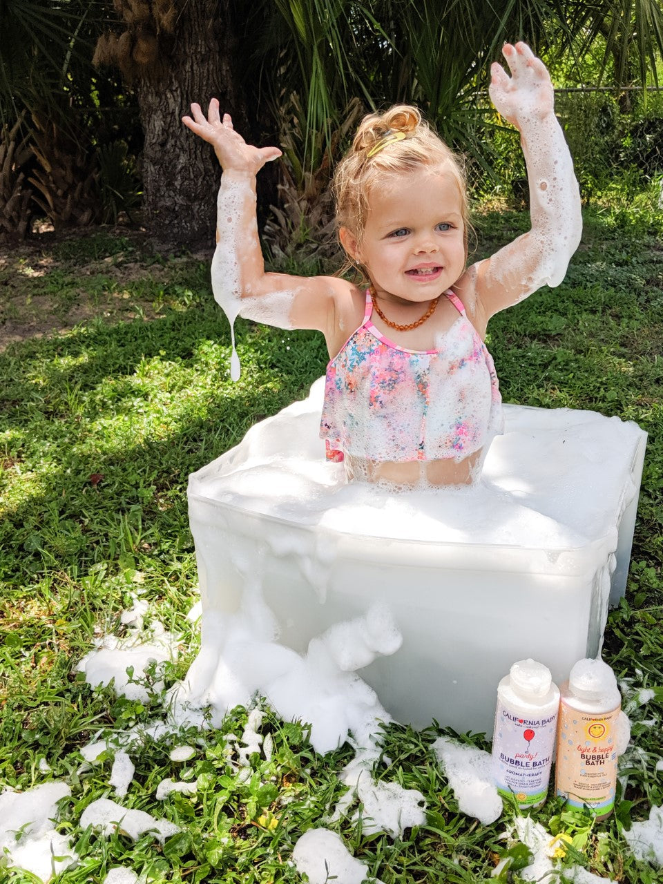 Child playing outdoors in a foam-filled tub surrounded by bubbles, showcasing the California Baby - Bubble Bath Fun Trio in a natural, playful setting.