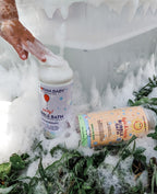 Foamy bath scene with two bottles of California Baby - Bubble Bath Fun Trio nestled among green leaves near a white bathtub, highlighting a playful and soothing bath time moment.