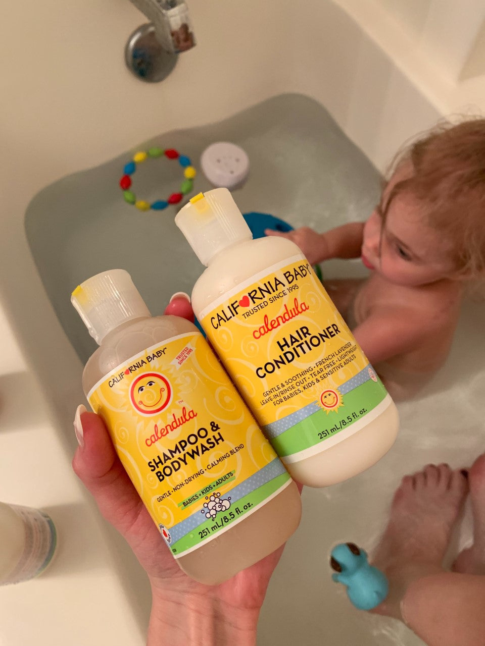Hand holding California Baby - Calendula™ Shampoo + Conditioner Bundle near a toddler bathing in a tub with bath toys, highlighting gentle, soothing baby hair and body care essentials.